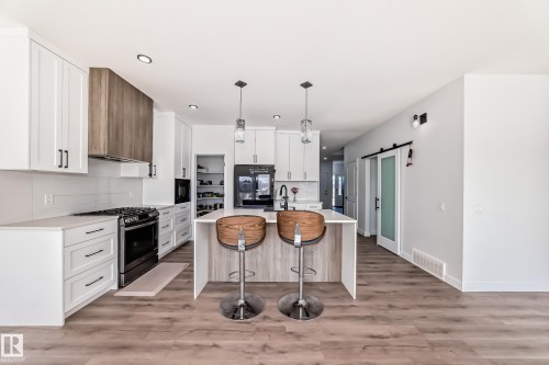 Modern kitchen featuring a central island with countertop seating, white cabinetry, integrated stainless steel appliances, a wood-finish range hood, and wood-finish flooring - 3247 Chernowski Way, Edmonton, AB - Indoor Photo Showing Kitchen With Upgraded Kitchen