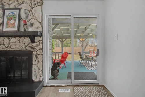 Sliding glass door opening to an outdoor patio with a pergola, complemented by a stone-faced fireplace with a dark mantel and hearth, and wood-finish flooring - 17305 60 Ave, Edmonton, AB 