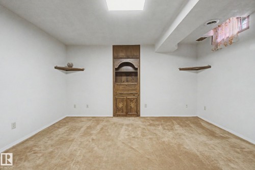 Carpeted room featuring a built-in wooden cabinet with shelving, two corner wall shelves, a ceiling-mounted light fixture, and a window with a valance - 17305 60 Ave, Edmonton, AB 