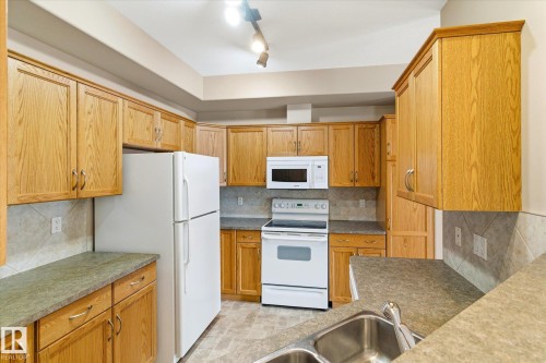 The kitchen features wood cabinetry, a double basin sink, and white appliances including a refrigerator, oven, and microwave - 104 612 111 Street Sw, Edmonton, AB - Indoor Photo Showing Kitchen