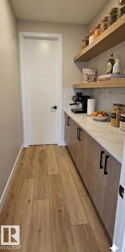 Dedicated pantry featuring wood-finish flooring, a white paneled door, built-in cabinetry with matte black hardware, white stone countertop, and two substantial wood floating shelves - 53 Mockingbird Cres, Fort Saskatchewan, AB - Indoor