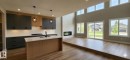 Open-concept living space featuring a modern kitchen island with a white countertop, two-tone cabinetry, wood-finish flooring, a contemporary fireplace, and abundant natural light through multiple windows - 53 Mockingbird Cres, Fort Saskatchewan, AB  - Indoor Photo Showing Kitchen With Fireplace 