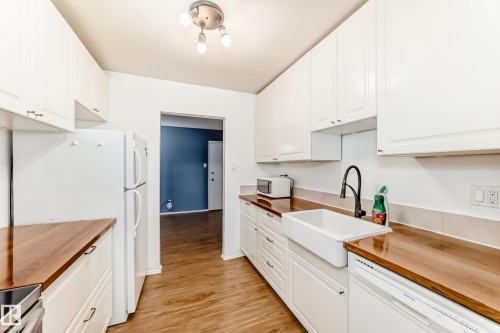 Kitchen featuring white cabinetry, wood-finish countertops, wood-finish flooring, a farmhouse-style sink with a matte black faucet, and white appliances - 423 5125 Riverbend Road, Edmonton, AB - Indoor Photo Showing Kitchen