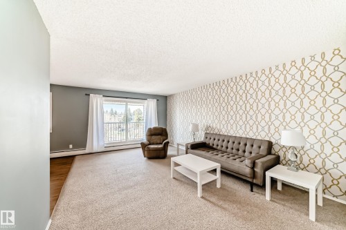 Spacious living area featuring a large window with white vertical blinds, patterned accent wall, and textured carpeting - 423 5125 Riverbend Road, Edmonton, AB - Indoor