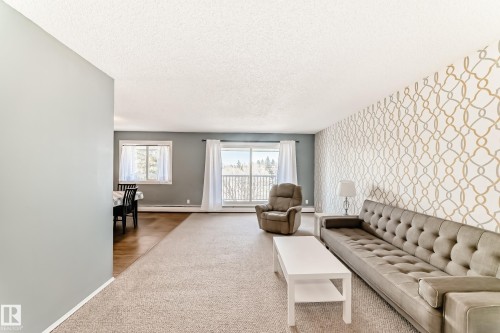 Spacious living area featuring patterned accent wall, light neutral carpeting, and a large window - 423 5125 Riverbend Road, Edmonton, AB - Indoor
