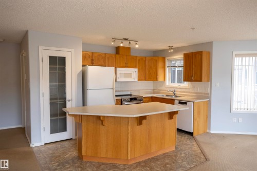 Kitchen featuring a central island with countertop, wood cabinetry, and white appliances - 101 2420 108 Street, Edmonton, AB - Indoor Photo Showing Kitchen With Double Sink