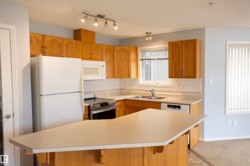 Kitchen featuring wood-finish cabinetry, white appliances, and a central island with an extended countertop - 101 2420 108 Street, Edmonton, AB - Indoor Photo Showing Kitchen With Double Sink