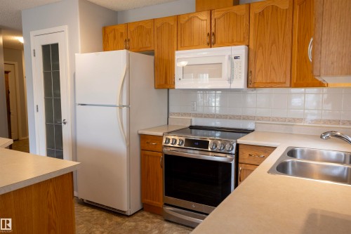 Kitchen featuring wood cabinetry, white countertops, stainless steel range, white over-range microwave, and a double basin stainless steel sink - 101 2420 108 Street, Edmonton, AB - Indoor Photo Showing Kitchen With Double Sink