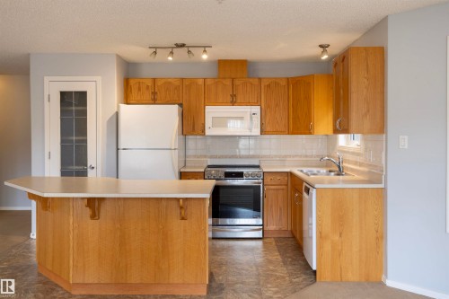 Kitchen with wood-finish cabinetry, a peninsula with countertop seating, white appliances, and track lighting - 101 2420 108 Street, Edmonton, AB - Indoor Photo Showing Kitchen