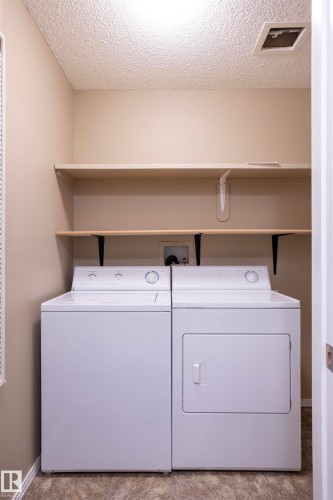 Dedicated laundry area featuring a white washing machine and dryer, two integrated storage shelves, and durable vinyl flooring - 101 2420 108 Street, Edmonton, AB - Indoor Photo Showing Laundry Room
