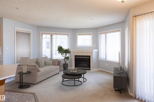 Living area featuring a corner fireplace with a tiled surround and mantel, multiple windows with vertical blinds, and light-colored carpeting - 101 2420 108 Street, Edmonton, AB - Indoor Photo Showing Living Room With Fireplace