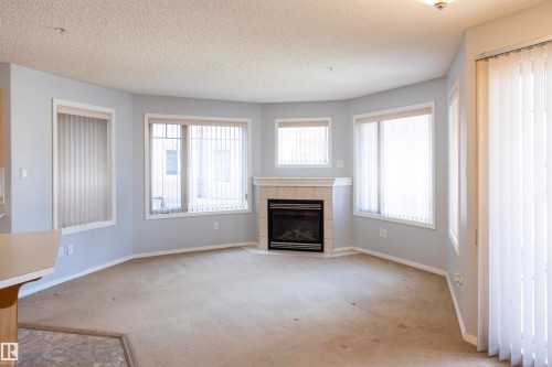 Living space featuring a corner fireplace with a tile surround and mantel, multiple windows with vertical blinds, and a light-colored carpet - 101 2420 108 Street, Edmonton, AB - Indoor Photo Showing Living Room With Fireplace