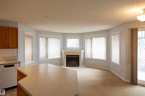 Open-concept living space featuring a fireplace with a light-toned surround, multiple windows with vertical blinds, a sliding glass door, and a kitchen island with a light-toned countertop - 101 2420 108 Street, Edmonton, AB - Indoor Photo Showing Other Room With Fireplace