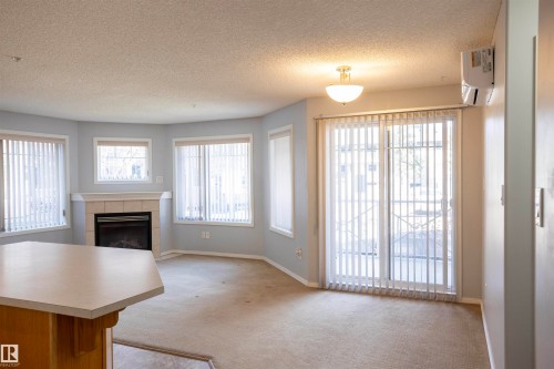Living area featuring a fireplace with a light-toned tile surround and mantle, multiple windows with vertical blinds, a ceiling-mounted light fixture, and a built-in air conditioning unit - 101 2420 108 Street, Edmonton, AB - Indoor Photo Showing Living Room With Fireplace