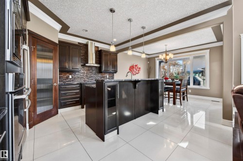 Kitchen featuring white tile flooring, dark wood cabinetry, a stone backsplash, stainless steel appliances, and a central island with a sink and pendant lighting - 1254 Adamson Drive, Edmonton, AB - Indoor Photo Showing Kitchen With Stainless Steel Kitchen With Upgraded Kitchen
