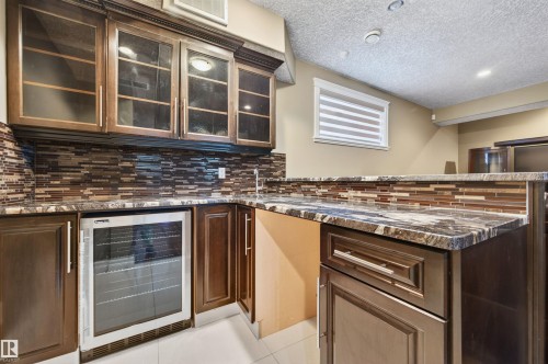 Built-in wet bar featuring a beverage cooler, rich wood cabinetry, glass-front upper cabinets, a mosaic tile backsplash, and stone countertops - 1254 Adamson Drive, Edmonton, AB - Indoor