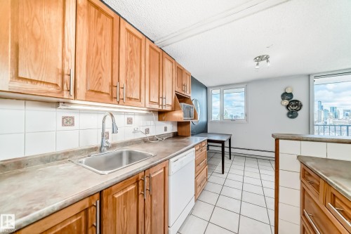 Kitchen featuring wood cabinetry, a stainless steel sink with a gooseneck faucet, a white tile backsplash with decorative accents, and tiled flooring - 405 10135 Saskatchewan Drive Nw, Edmonton, AB - Indoor Photo Showing Kitchen