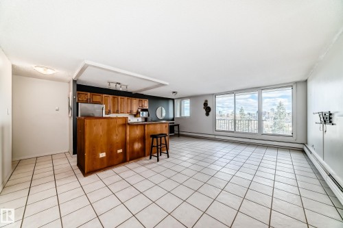 Open-concept living area featuring large tile flooring, a kitchen with wood cabinetry and stainless steel appliances, and a wall of windows overlooking a cityscape - 405 10135 Saskatchewan Drive Nw, Edmonton, AB - Indoor