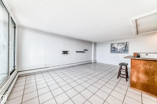 Bright open concept room featuring large format tile flooring, expansive windows, a wood-finish bar, and a wall-mounted display bracket - 405 10135 Saskatchewan Drive Nw, Edmonton, AB - Indoor Photo Showing Other Room
