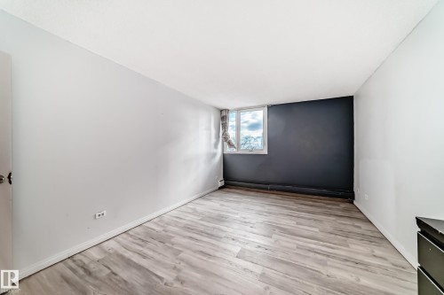 Light wood-finish flooring provides a neutral base for this room featuring a window with white trim, a dark accent wall, and white baseboards - 405 10135 Saskatchewan Drive Nw, Edmonton, AB - Indoor Photo Showing Other Room