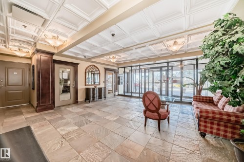 Foyer featuring natural stone tile flooring, coffered ceilings with integrated lighting, glass entry doors, a dark wood-paneled column, and an arched mirror with a console table - 405 10135 Saskatchewan Drive Nw, Edmonton, AB - Indoor Photo Showing Other Room