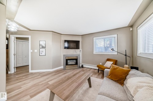 Living area featuring light wood-finish flooring, a built-in fireplace with a stone-finish hearth, multiple windows with blinds, and white baseboards - 101 10827 85 Avenue Nw, Edmonton, AB - Indoor Photo Showing Living Room With Fireplace