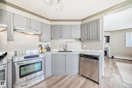 Contemporary kitchen featuring light gray cabinetry, stainless steel appliances, marble-look countertops, and wood-finish flooring - 101 10827 85 Avenue Nw, Edmonton, AB - Indoor Photo Showing Kitchen