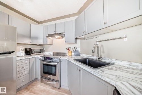Contemporary kitchen featuring light gray cabinetry, stainless steel appliances, a black undermount sink, and white marble-patterned countertops - 101 10827 85 Avenue Nw, Edmonton, AB - Indoor Photo Showing Kitchen