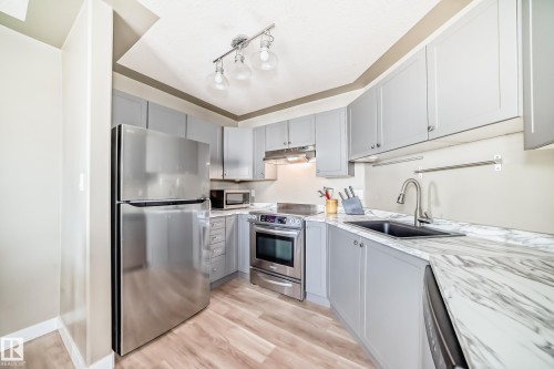 Modern kitchen featuring light gray cabinetry, stainless steel appliances, marble-patterned countertops, a stainless steel sink with a gooseneck faucet, and wood-finish flooring - 101 10827 85 Avenue Nw, Edmonton, AB - Indoor Photo Showing Kitchen