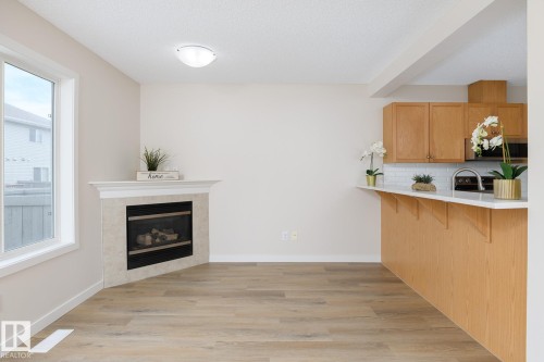 Spacious open-concept area featuring wood-finish flooring, a corner fireplace with a white mantel and stone surround, and abundant natural light from a large window - 55 14603 Miller Boulevard, Edmonton, AB - Indoor Photo Showing Other Room With Fireplace