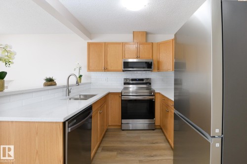 U-shaped kitchen featuring light wood cabinetry, stainless steel appliances, a subway tile backsplash, white countertops, and wood-finish flooring - 55 14603 Miller Boulevard, Edmonton, AB - Indoor Photo Showing Kitchen With Stainless Steel Kitchen With Double Sink