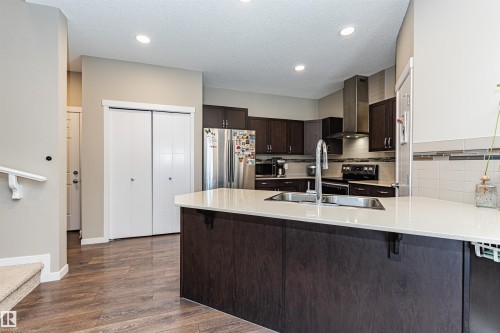 Kitchen featuring an island with a built-in sink, stainless steel appliances, dark wood-finish cabinetry, light-toned countertops, and a subway tile backsplash - 4452 Annett Common, Edmonton, AB 