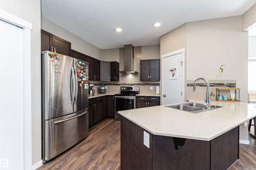 Kitchen featuring a substantial island with a double basin sink and a chrome faucet, dark wood-finish cabinetry, stainless steel appliances, recessed lighting, and wood-finish flooring - 4452 Annett Common, Edmonton, AB 
