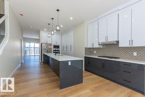 Contemporary kitchen featuring wood-finish flooring, two-tone cabinetry with white upper and dark lower units, an elongated center island with pendant lighting, a tile backsplash, and stainless steel appliances - 9814 155 Street, Edmonton, AB - Indoor Photo Showing Kitchen With Upgraded Kitchen