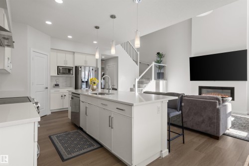 Open-concept living area featuring a kitchen island with seating, stainless steel appliances, wood-finish flooring, a contemporary fireplace, and a staircase with a white railing - 1471 Plum Circle Circle, Edmonton, AB - Indoor Photo Showing Kitchen With Upgraded Kitchen