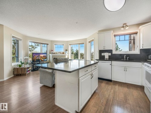 Open-concept living area featuring a kitchen island with dark countertops, white cabinetry, integrated appliances, and wood-finish flooring throughout - 317 2420 108 Street, Edmonton, AB - Indoor Photo Showing Kitchen