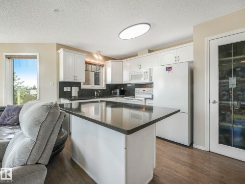 Bright kitchen featuring white cabinetry, dark countertops, white appliances, a central island, and wood-finish flooring - 317 2420 108 Street, Edmonton, AB - Indoor Photo Showing Kitchen