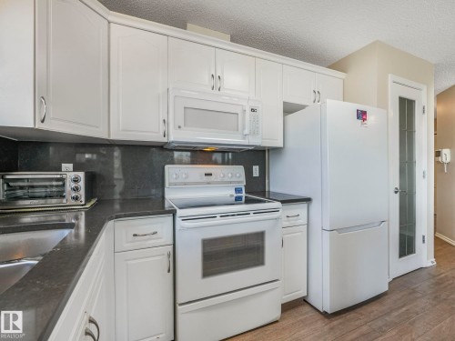 Kitchen featuring white cabinetry with brushed nickel hardware, a white range, a white microwave, a white refrigerator, and dark countertops with a coordinating backsplash - 317 2420 108 Street, Edmonton, AB - Indoor Photo Showing Kitchen With Double Sink