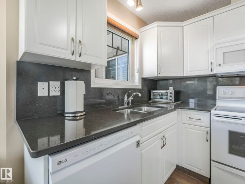 Kitchen featuring white cabinetry with silver hardware, dark countertops, an undermount double basin sink with gooseneck faucet, a built-in dishwasher, and a white oven - 317 2420 108 Street, Edmonton, AB - Indoor Photo Showing Kitchen With Double Sink