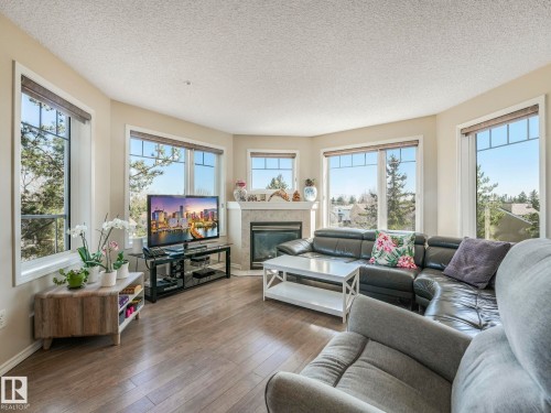 Living area with wood-finish flooring, a corner fireplace featuring a stone surround and mantel, and multiple large windows with trim - 317 2420 108 Street, Edmonton, AB - Indoor Photo Showing Living Room With Fireplace