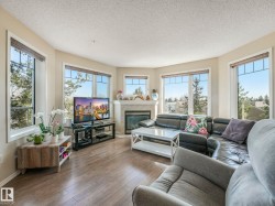 Living area with wood-finish flooring, a corner fireplace featuring a stone surround and mantel, and multiple large windows with trim - 