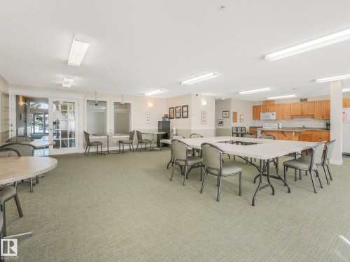 Spacious common area featuring textured carpeting, multiple tables with seating, a built-in kitchen with wood-finish cabinetry, and recessed fluorescent lighting - 317 2420 108 Street, Edmonton, AB - Indoor