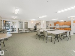 Spacious common area featuring textured carpeting, multiple tables with seating, a built-in kitchen with wood-finish cabinetry, and recessed fluorescent lighting - 