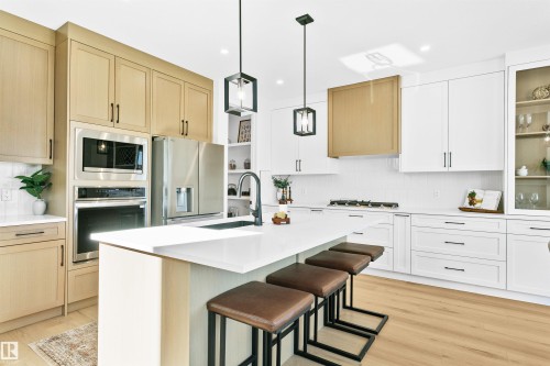 Modern kitchen featuring a central island with white countertop, light wood-finish flooring, white flat-panel cabinetry, and integrated stainless steel appliances - 34 Harley Way, Spruce Grove, AB - Indoor Photo Showing Kitchen With Upgraded Kitchen