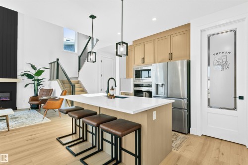 Contemporary open-concept kitchen and living area featuring an expansive island with seating, a full suite of stainless steel appliances, light wood-finish flooring, a modern fireplace with a dark paneled accent wall, and a staircase with dark railings - 34 Harley Way, Spruce Grove, AB - Indoor Photo Showing Kitchen With Upgraded Kitchen