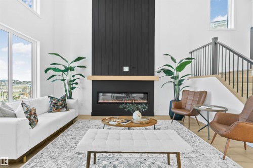 Double-height living space featuring large windows, wood-finish flooring, a contemporary linear fireplace with a black paneled surround, and an open staircase with black iron balusters - 34 Harley Way, Spruce Grove, AB - Indoor Photo Showing Living Room With Fireplace