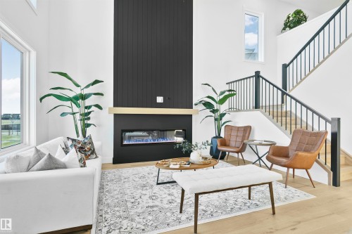 Spacious living area featuring high ceilings, an electric linear fireplace with a dark paneled surround and light wood mantel, a multi-level staircase with dark banisters, and large windows providing natural light - 34 Harley Way, Spruce Grove, AB - Indoor Photo Showing Living Room With Fireplace