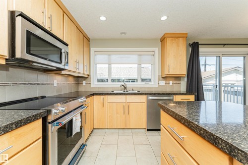 Kitchen featuring light wood cabinetry, dark granite-look countertops, stainless steel appliances, a tile backsplash, and ceramic tile flooring - 8530 76 Avenue, Edmonton, AB - Indoor Photo Showing Kitchen With Double Sink