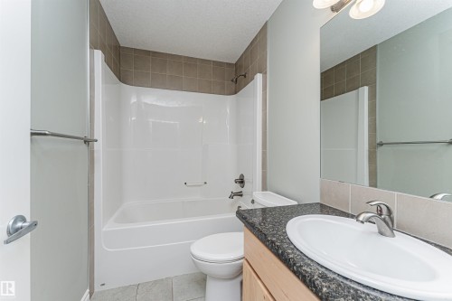Bathroom featuring a white bathtub and shower combination with taupe tile surround, a light wood-finish vanity with a dark speckled countertop, an undermount white sink, and a large wall-mounted mirror - 8530 76 Avenue, Edmonton, AB - Indoor Photo Showing Bathroom