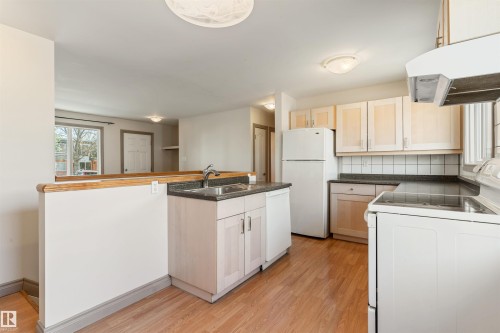 Functional kitchen featuring wood-finish flooring, light wood cabinetry, white appliances, and dark speckled countertops - 10620 68 Avenue, Edmonton, AB - Indoor Photo Showing Kitchen With Double Sink
